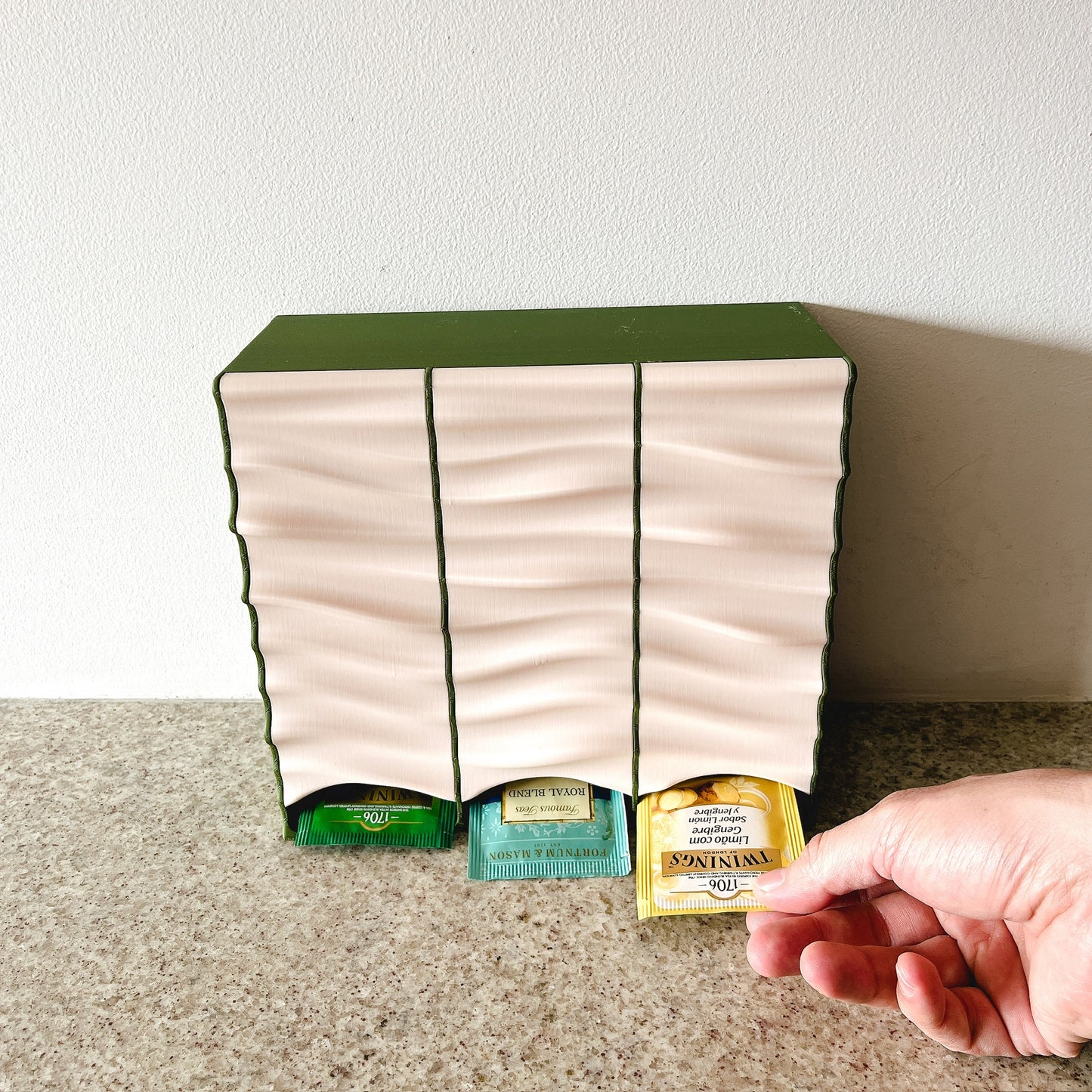 Dispensador Verde oliva + Beige, mano sacando bolsita por el frente, mostrando uso real
