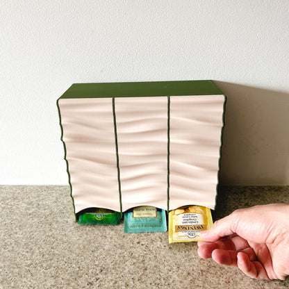 Dispensador Verde oliva + Beige, mano sacando bolsita por el frente, mostrando uso real