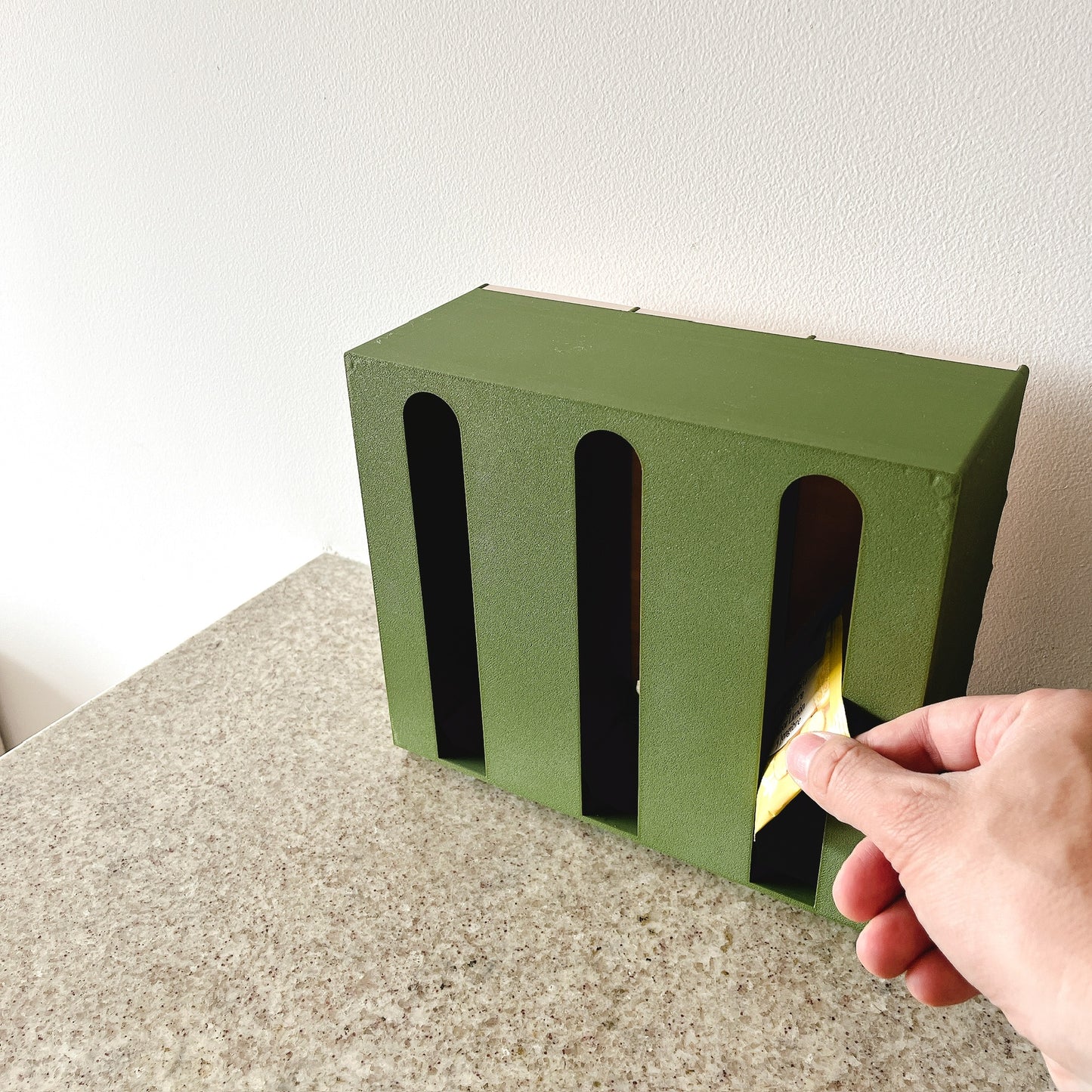 Dispensador Verde oliva + Beige, mano sacando bolsita por las aberturas en arco del lado opuesto