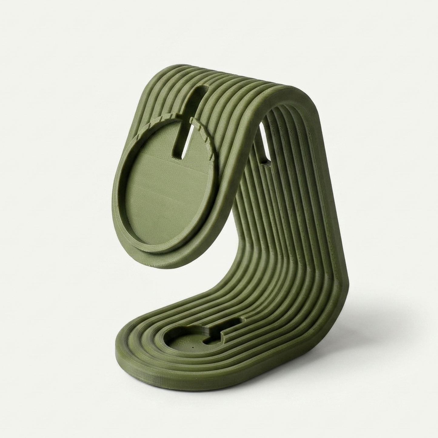 Soporte de carga matte dark green para iPhone y Apple Watch, vista lateral 3/4 mostrando el perfil curvado, sobre mesa de madera, fondo de pared blanca