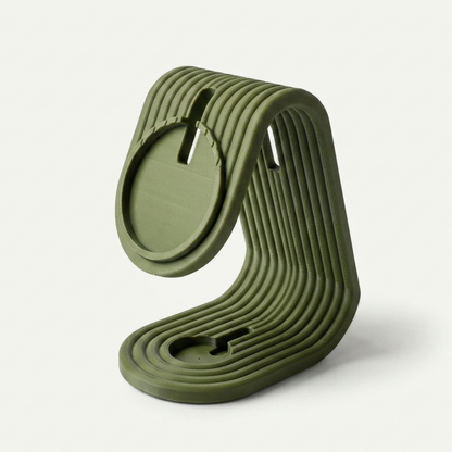 Soporte de carga matte dark green para iPhone y Apple Watch, vista lateral 3/4 mostrando el perfil curvado, sobre mesa de madera, fondo de pared blanca